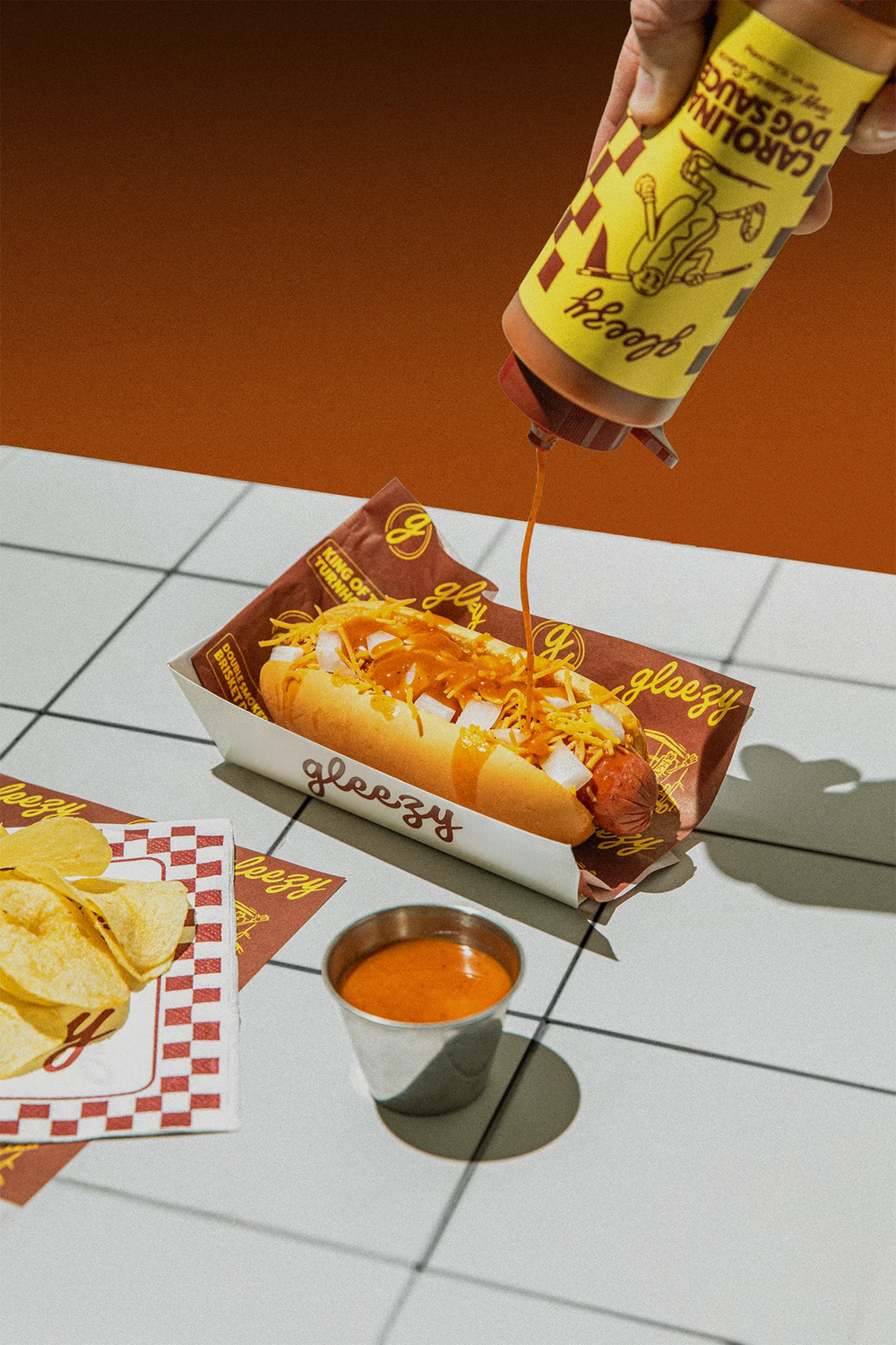 A Gleezy Carolina Dog Sauce bottle sitting next to two cheese dogs sitting on Gleezy branded paper boxes.