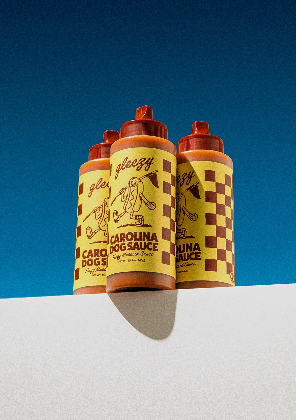 A Gleezy Carolina Dog Sauce bottle sitting next to two cheese dogs sitting on Gleezy branded paper boxes.
