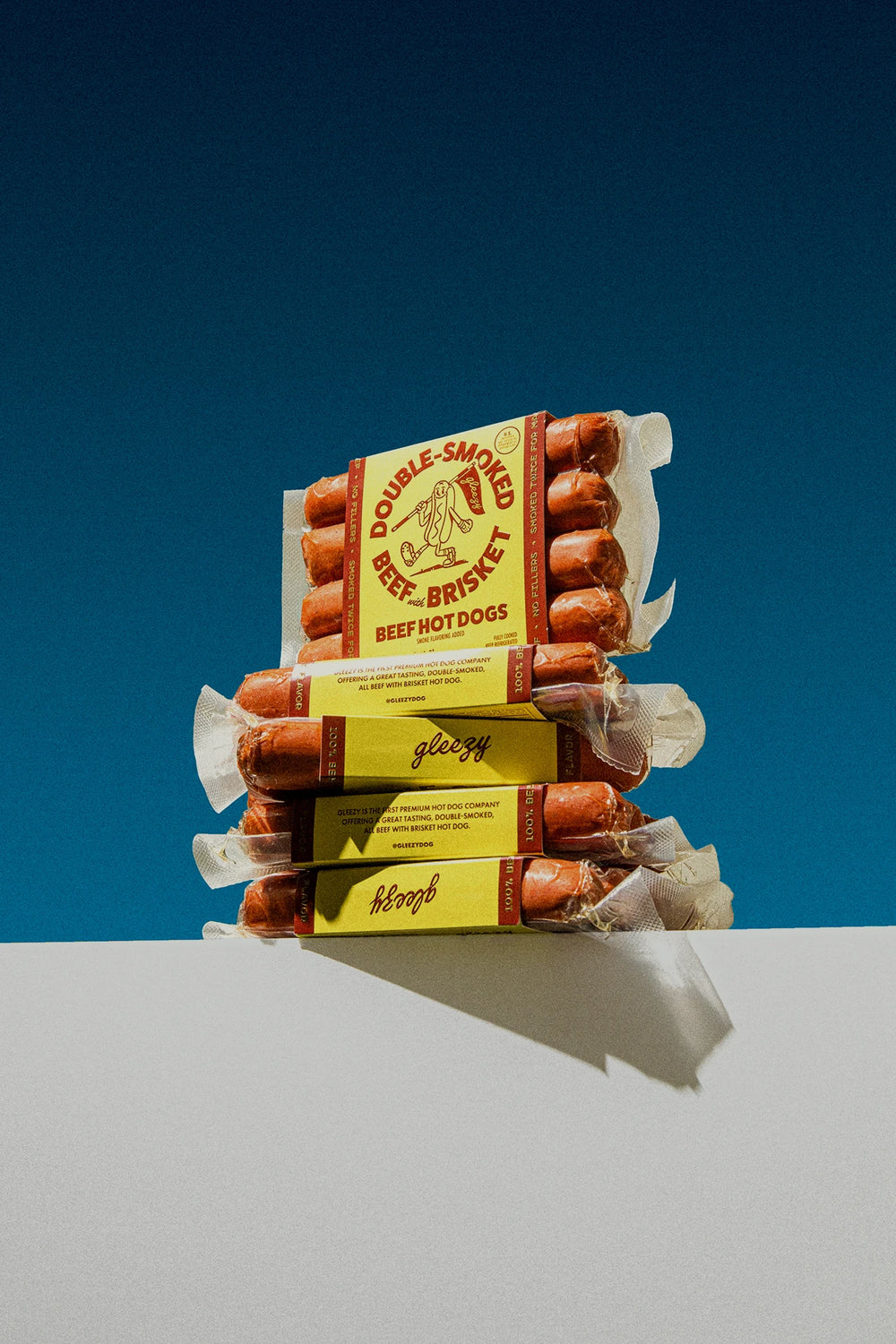 A Gleezy Carolina Dog Sauce bottle sitting next to two cheese dogs sitting on Gleezy branded paper boxes.