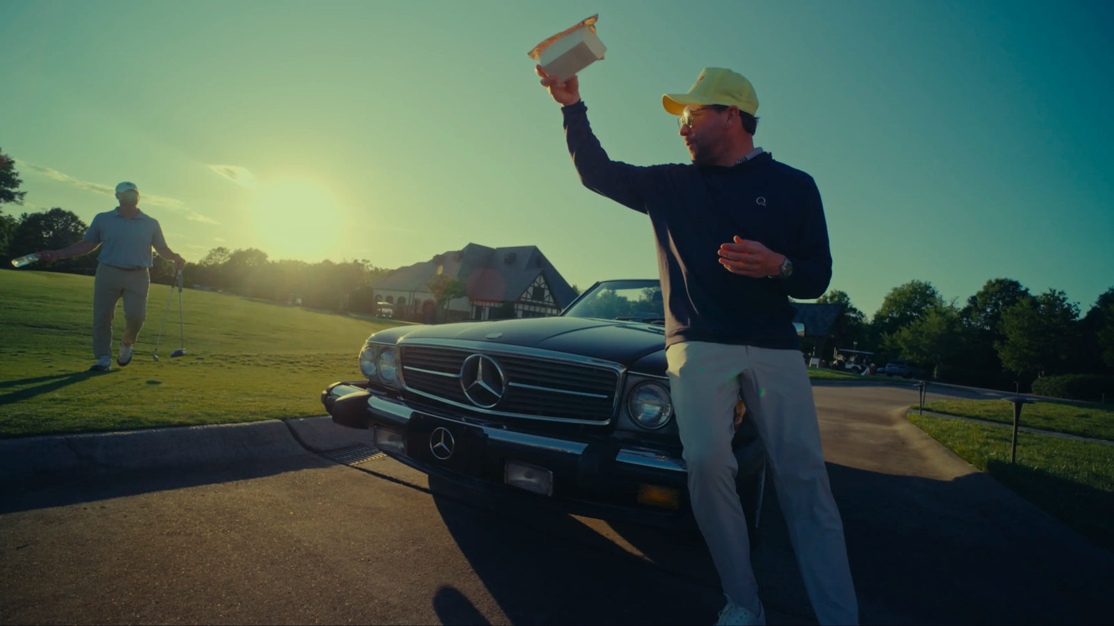 Man in golf attire holding up a Gleezy King of the Box game while leaning on a vintage Mercedes convertible at a golf course
