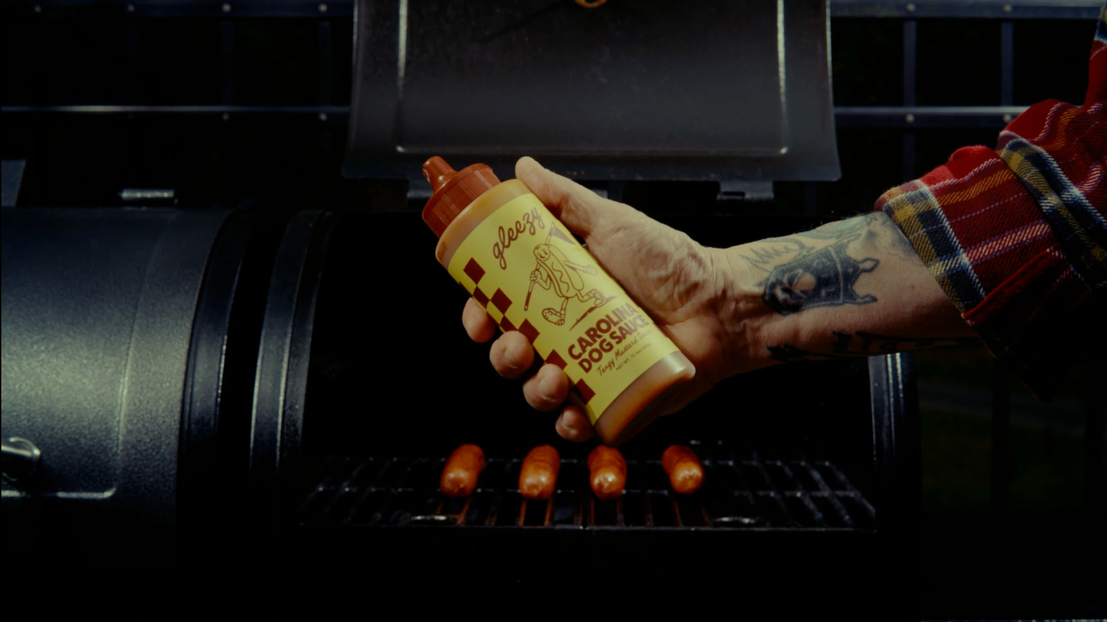 A person holding a bottle of Gleezy Carolina Dog Sauce in front of an open bbq grill.